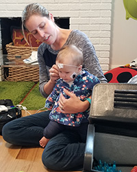 A blonde-haired woman wearing blue jeans and kneeling on a laminate floor holds an infant with a white lead attached to its head.