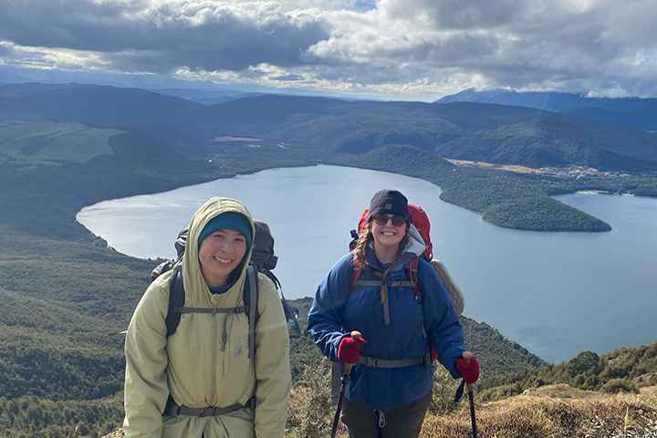 Two young women wearing rain gear pose for a photo with a lake, lakeshore and partly cloudy sky in the background.