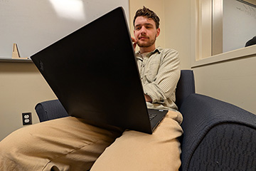A young man with facial hair and wearing a button down shirt and khakis looks at a laptop screen.
