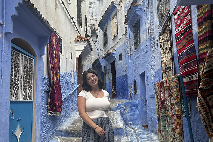 A young smiling woman with brown hair and wearing a white top stands on a cobblestone road bordered with blue and white brick buildings decorated with multi-colored rugs.