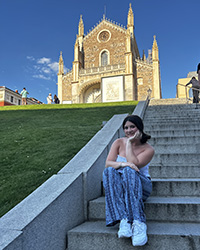 A young woman in multi-hued pants sits on an outdoor staircase with an old church in the background.