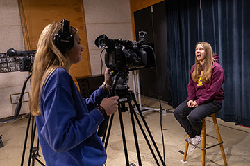 A woman in a maroon sweatshirt sitting on a stool talks while a young woman with long blonde hair and a blue sweatshirt records her on a video camera.