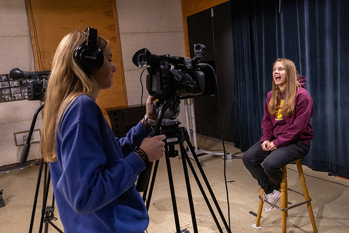 A woman in a maroon sweatshirt sitting on a stool talks while a young woman with long blonde hair and a blue sweatshirt records her on a video camera.