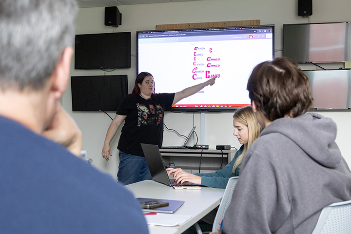 A young woman in a black T-shirt points to a large television screen with the word CAMEO written on it in several different fonts while a gray-haired man a young man in a gray hoodie look on and a young woman in a glue-grey top looks at a laptop screen.