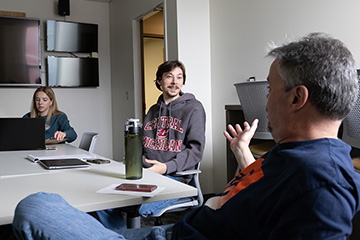 A man with gray hair and a blue shirt gestures while talking across a white table to a young man in a gray Central Michigan sweatshirt while a woman in a blue-green top looks at a laptop screen.