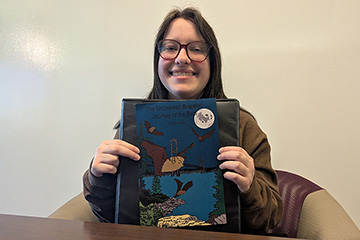 A smiling woman with long brown hair parted in the middle holds up a book with an image of a bat on the cover