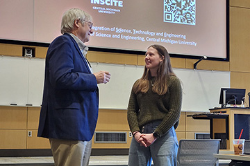 A man with gray hair and glasses and wearing a blue suitcoat talks to a young woman with long hair and a sweater in front of a screen with Integration of Science Technology and Engineering written on it.