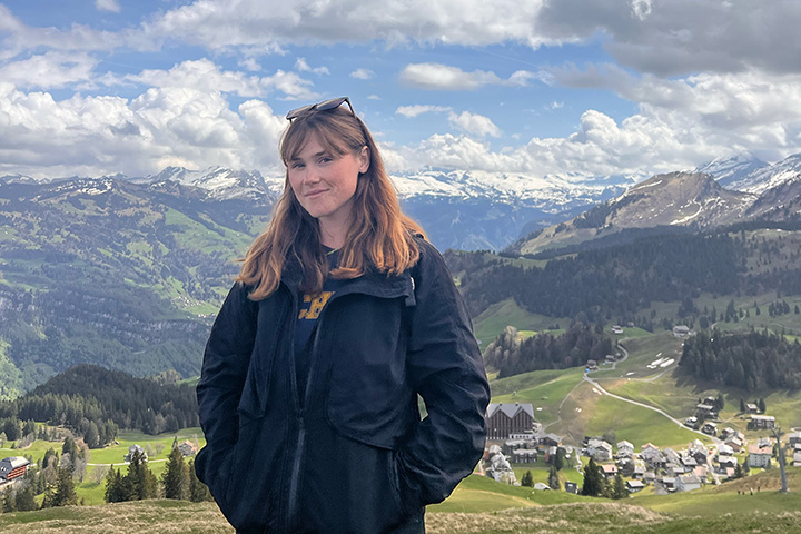 A smiling woman with long red hair with sunglasses perched atop her head has her hands in a zipped coat with a hilly landscape in the background