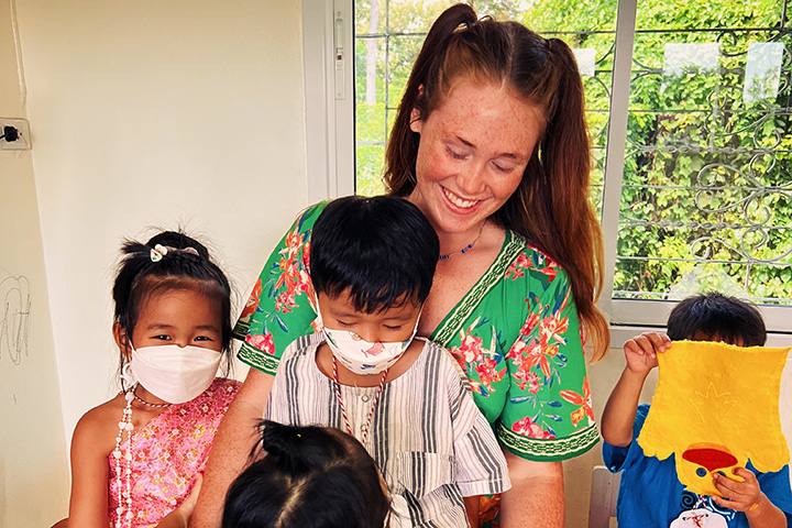 A smiling woman with red hair and a printed top looks down at three masked children.