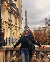 A woman with long red hair and black sunglasses and wearing a black leather jacket leans against a railing with the Eifel Tower in the background