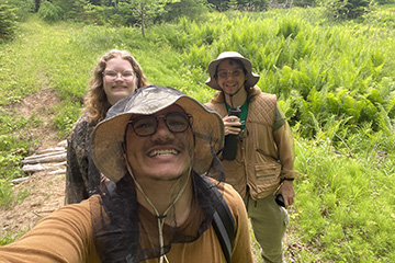 A smiling young man wearing a boonie hat topped with mosquito netting takes a selfie pic with a smiling young woman with curly hair and another young man who is also wearing a boonie hat stand behind him