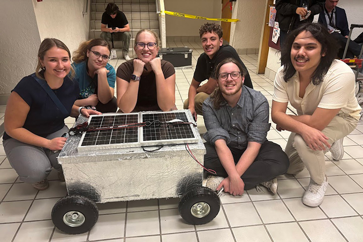 Six young people--three men and three women--pose for a photo while crouched and sitting behind a shiny metal box on wheels