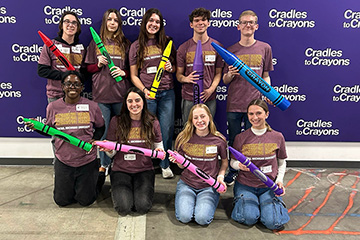 Nine young people wearing maroon and gold t shirts with central michigan university written on them and holding oversized crayons post before a purple backdrop with cradles to crayons written on it multiple times