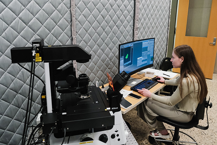 A woman with straight brown hair and wearing a beige dress adjusts images on a computer monitor with a microscope in the foreground