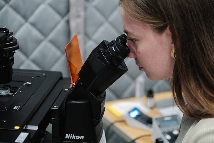 A woman with straight long brown hair looks into a microscope eyepiece