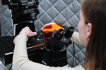 A woman with straight brown hair prepares a microscope sample behind an eyepiece and opaque orange shield