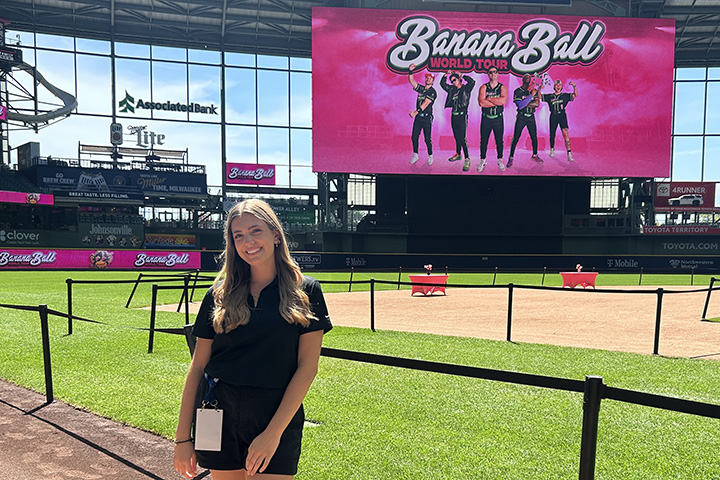 A young woman with long blonde hair parted in the center and wearing a maroon top leans against a railing with a baseball field in the background and a large sign that reads Banana Ball.