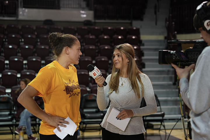 A young woman in a white sweater holds a microphone to a woman in a gold T shirt
