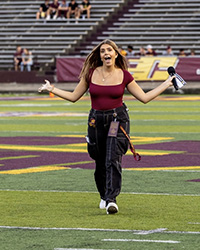 A young woman with long hair and wearing a maroon top and black denim pants walks across a football field towards a camera with outstretched hands holding a microphone