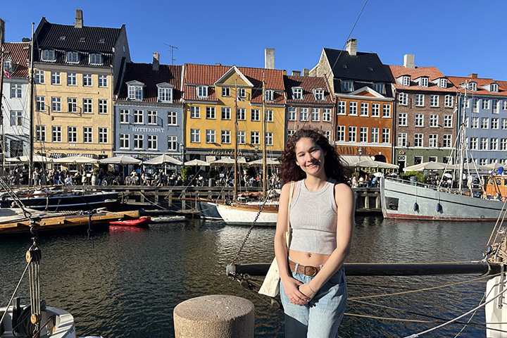 A young woman with curly hair and a gray top poses in front of a canal lined with different colored buildings