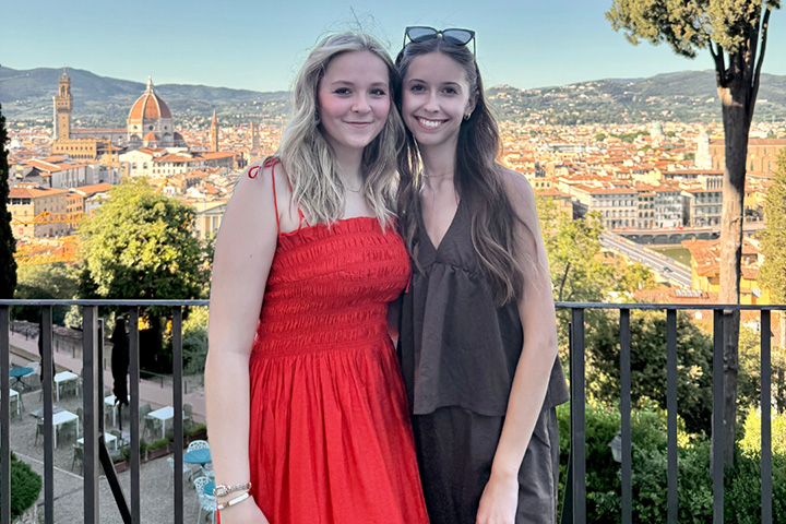 A woman with long blond hair and wearing a red dress poses with a young woman with long brown hair and a brown dress on a balcony overlooking a city