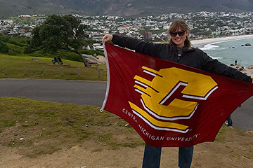 A woman with black sunglasses poses with a maroon flag with a large gold C on it in front of a city