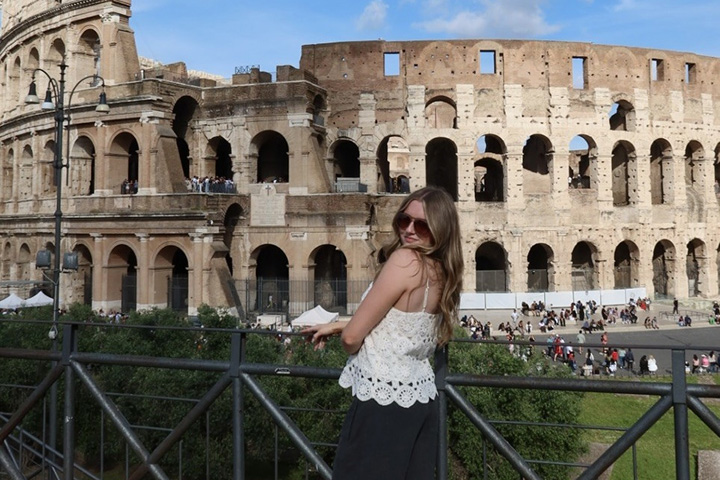 A young woman with long brown hair and black sunglasses stands next to a railing in front of a old collesium