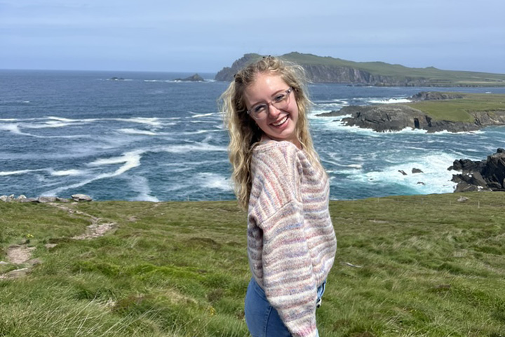 A young woman with long blonde hair and glasses stands on grass in front of a large body of water