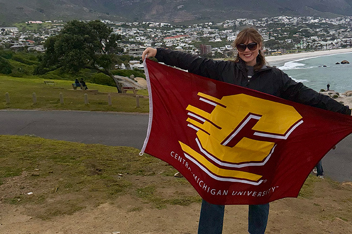 A woman with black sunglasses poses with a maroon flag with a large gold C on it in front of a city