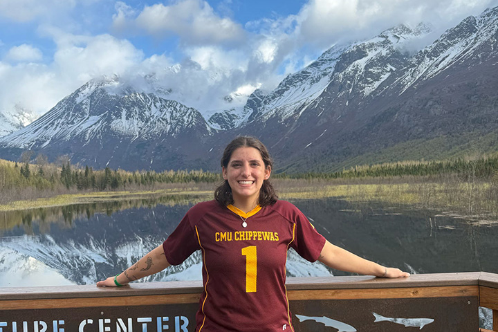 A woman wearing a maroon jersey with CMU Chippewas 1 on it stands on an overlook with cloud shrouded and snow capped mountains in the background