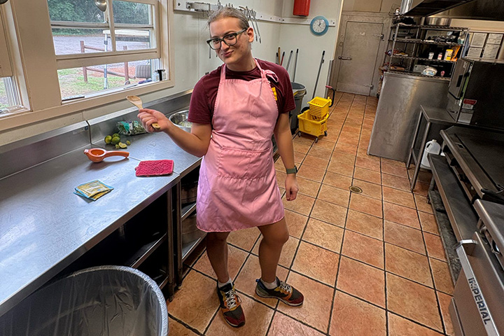 A young woman wearing a pink apron points with her thumb towards a metal food preparation counter