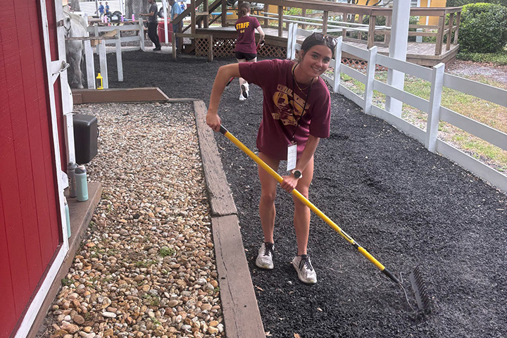 A young woman wearing a maroon T-shirt spreads black gravel on a driveway