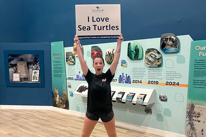 A young woman wearing a dark T-shirt and shorts holds a sign that reads I Love Sea Turtles
