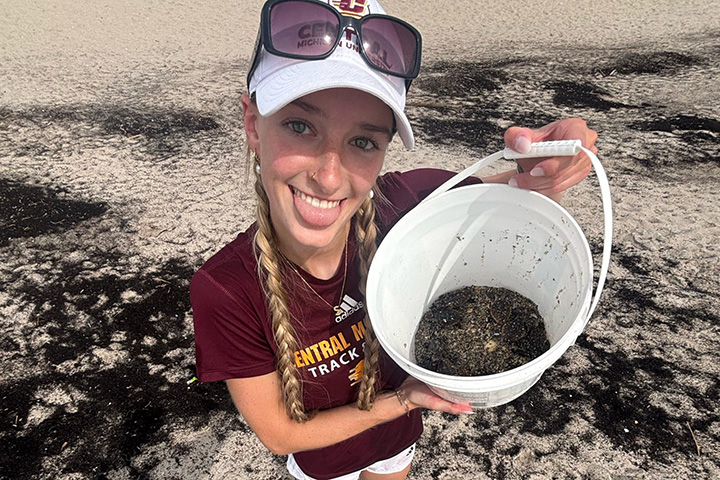 A woman with braided blonde hair and wearing a white baseball cap and sunglasses and sticking out her tongue poses with a bucket with dirt and a baby sea turtle at the bottom