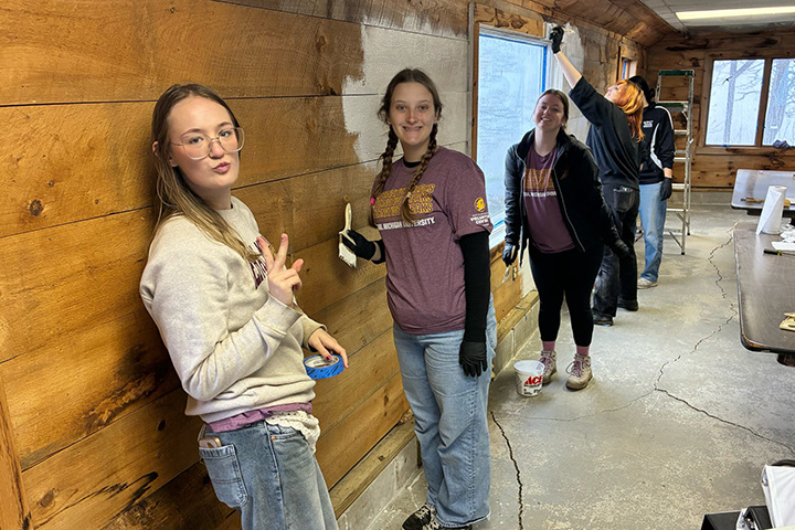 Two young women pose in front of a wooden plank wall while several other young people clean and repair the wall.