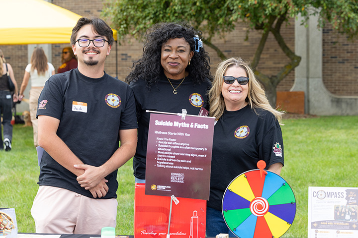 A man and two women in black tee shirts pose smiling outside and in front of a placard