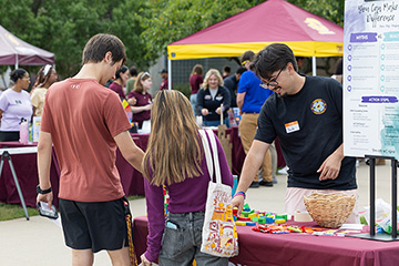A man with parted black hair and glasses wearing a black T-shirt points to a table with a maroon table cloth for the benefit of a man in black shorts and a rust colored tee shirt and a woman with long hair wearing blue jeans and a wine colored top