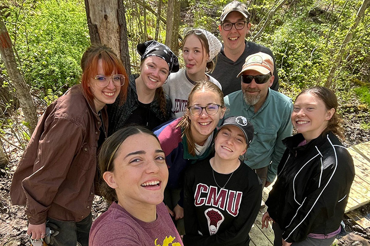 A group of nine people of various ages and genders smile while posing for a picture while standing on a boardwalk in the middle of the forest