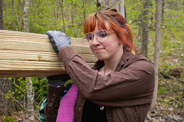A woman with copper colored hair and square rimmed glasses and wearing work gloves and a brown jacket holds four pieces of lumber on her right shoulder