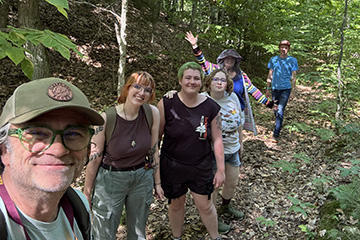 A man wearing glasses and a ball cap smiles while five other people are arrayed loosely in a line on a trail in the forest