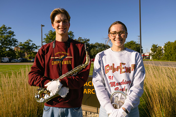 A young man in a maroon sweatshirt and white gloves and holding a saxophone stands next a young woman in a gray sweatshirt holding a horn in a field on a sunny day