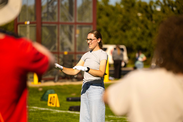 A young woman with long hair and glasses holds two gloved hands parallel to the ground while standing on a turf field