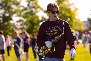 A young man wearing black sunglasses and a maroon sweatshirt holds a saxophone that hangs around his chest with a hand in a white glove