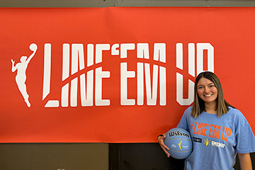 A young woman wearing a blue tee shirt smiles while standing in front of a banner that reads line em up