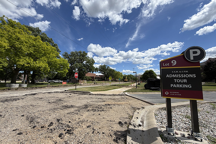 A street is torn up with construction vehicles in the background. In the foreground, a sign reads Admissions Tour Parking.