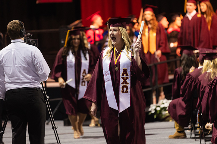 A blonde female graduate smiles at the crowd while holding her diploma
