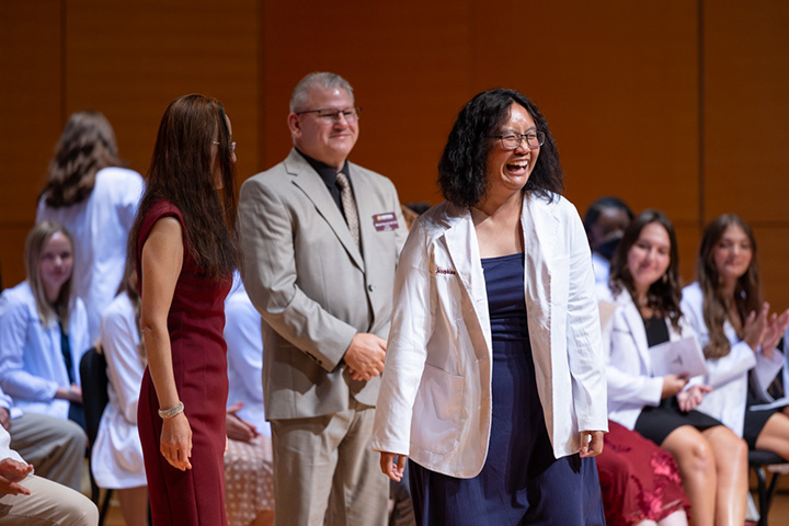 A physician assistant student smiles while standing on stage and receiving her white coat