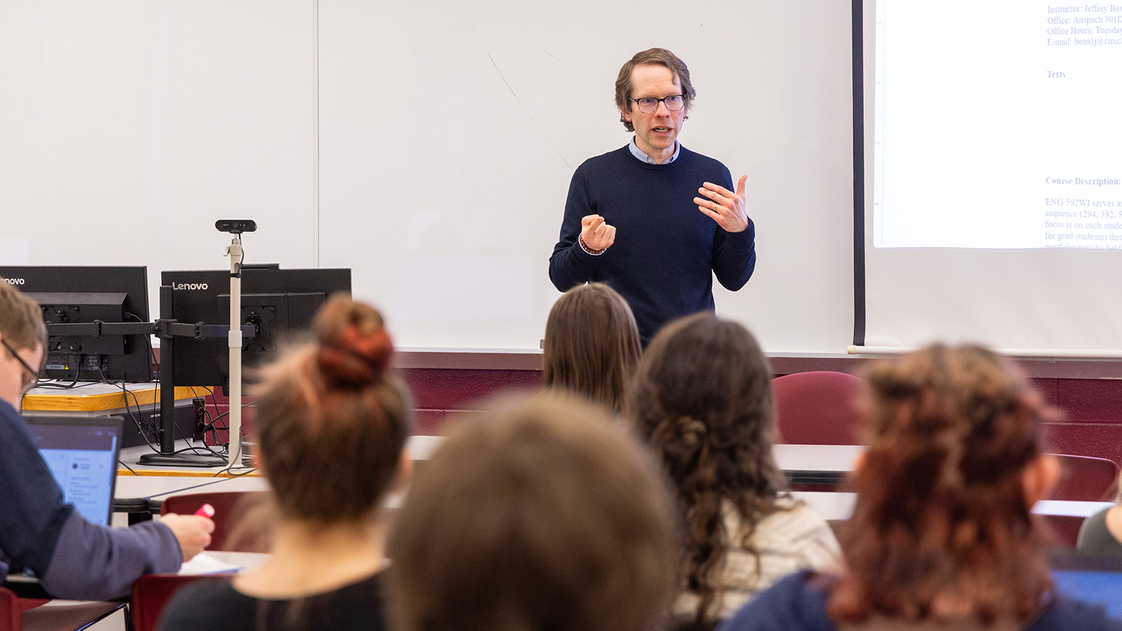 26-01-14 First Day of Classes - mbh -11-1600x900 A professor in a black sweater standing in front of a classroom full of students.