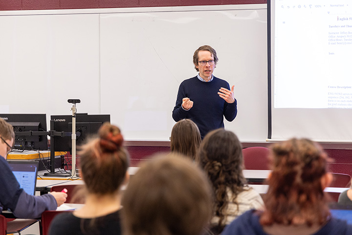 A professor standing in front of a whiteboard with a group of students in the background