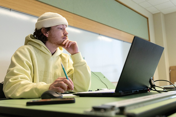 A male student in a beanie and a yellow sweatshirt looking at a laptop